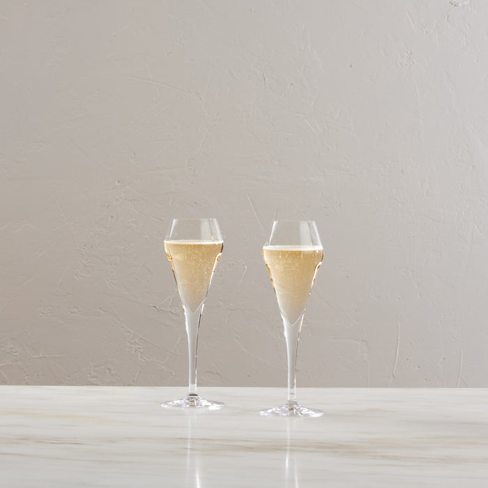 Two clear flute glasses filled with sparkling wine on a white surface.
