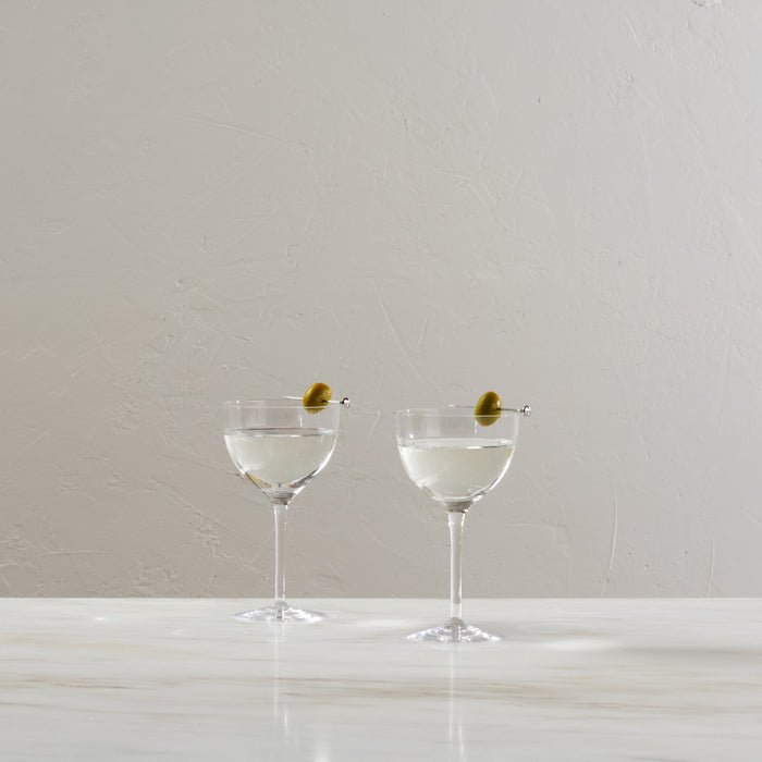 Two coupe glasses filled with clear liquid and garnished with green olives on picks.
