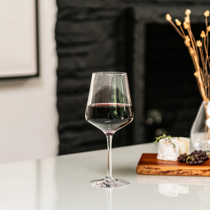 Red wine in a stemmed glass on white table with cheese, grapes, and decorative elements in background.