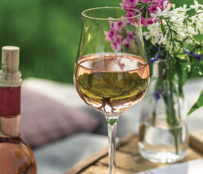 Tulip wine glass filled with white wine, surrounded by fresh flowers and wine bottles on outdoor table.