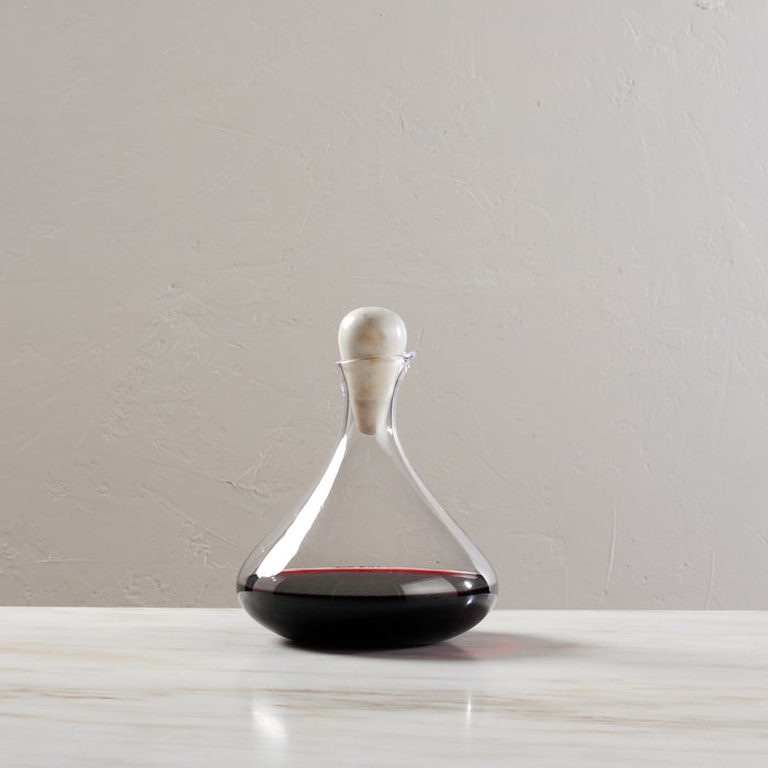 Clear glass wine decanter with wide base, tapered neck, and white marble spherical stopper, containing red wine.