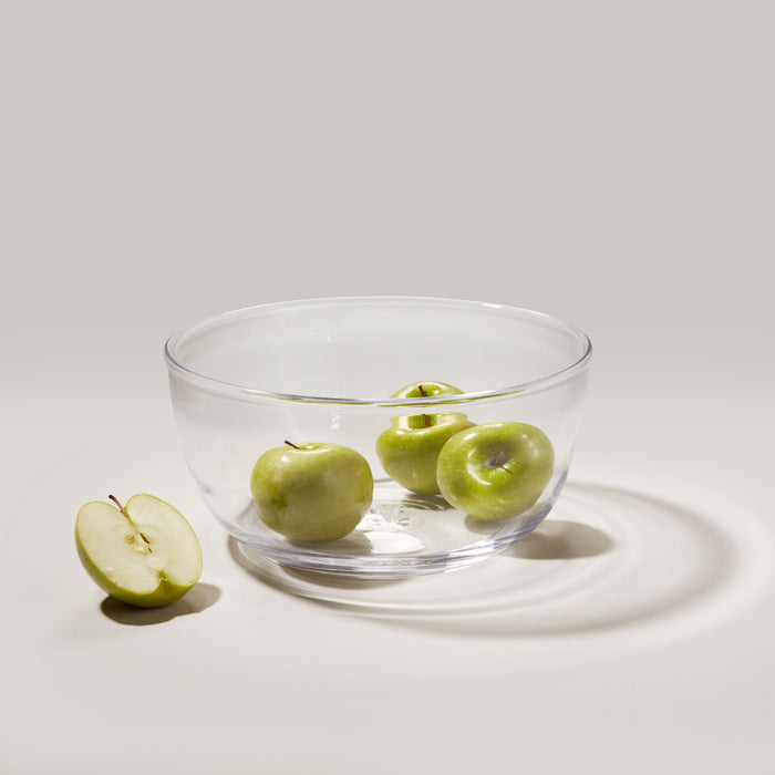 Large clear glass bowl with wide rim containing three green apples, one apple halved beside it.