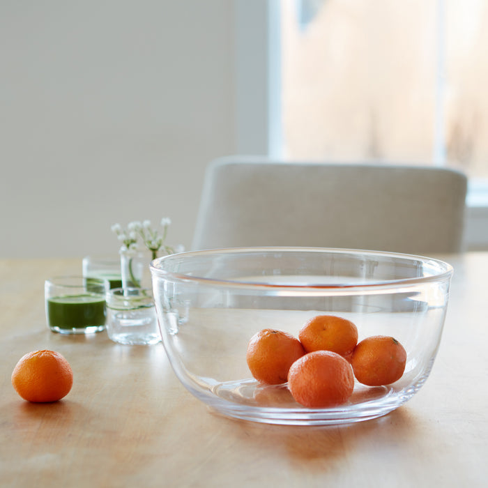 Clear glass bowl containing small oranges on a wooden table with glasses and flowers in background.