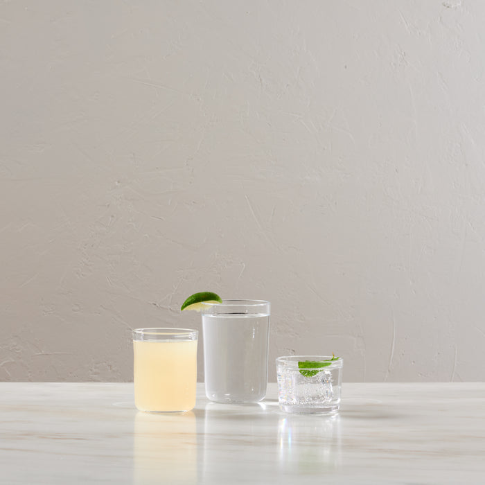 Three clear glass cups of varying heights containing different beverages with lime garnishes on a white surface.