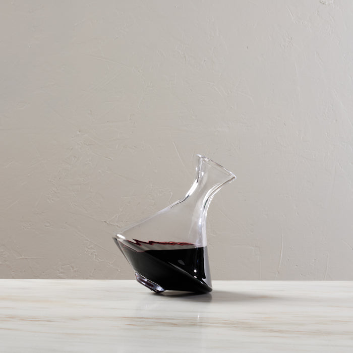 Clear glass wine decanter with curved spout, tilted base, partially filled with red wine.