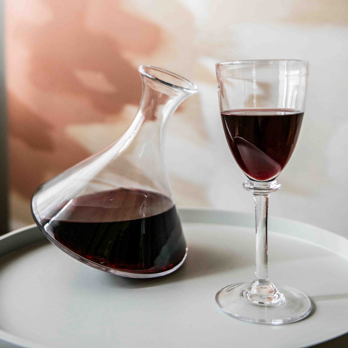 Glass wine decanter with curved spout filled with red wine, displayed next to a filled wine glass on a white plate.