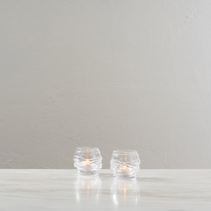 Two clear glass tealight holders with lit candles inside, placed on a white surface against a textured gray wall.