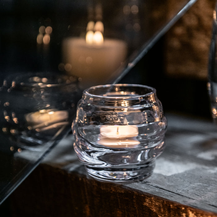 Lit tealight candle in clear glass holder with additional burning candles blurred in background.