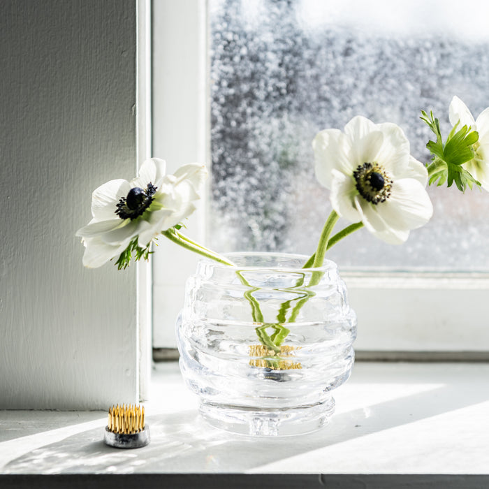 Two tealight candles with gold-colored bases beside a clear glass vase holding white anemone flowers on a windowsill.