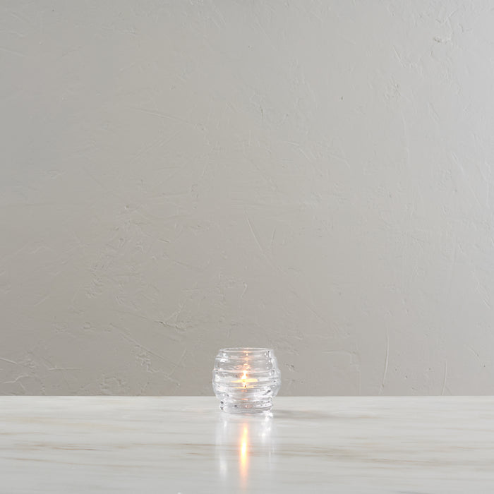 Clear glass tealight holder with lit candle inside, textured surface.