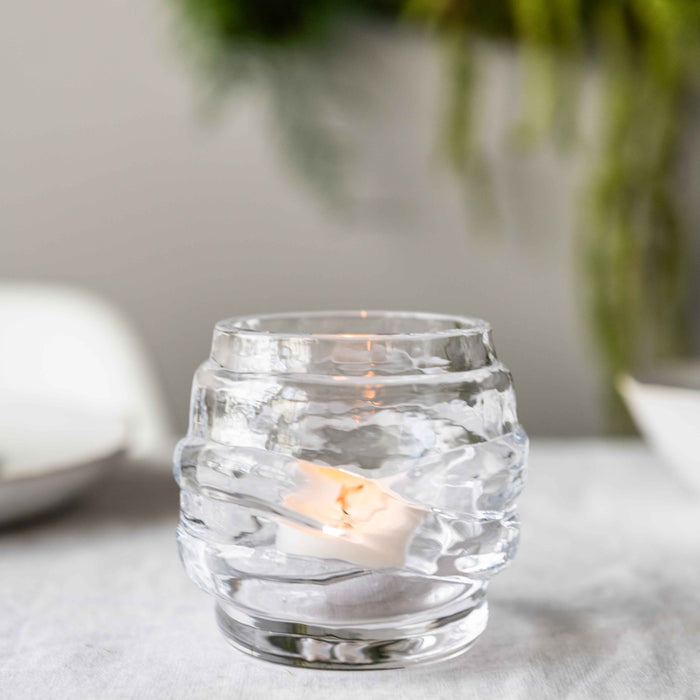 Clear glass tealight holder with horizontal ridged pattern and lit tealight inside.