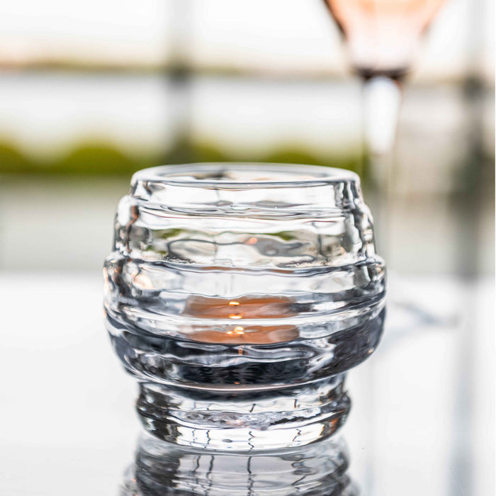Clear glass tealight holder with horizontal ridged texture and lit candle inside.