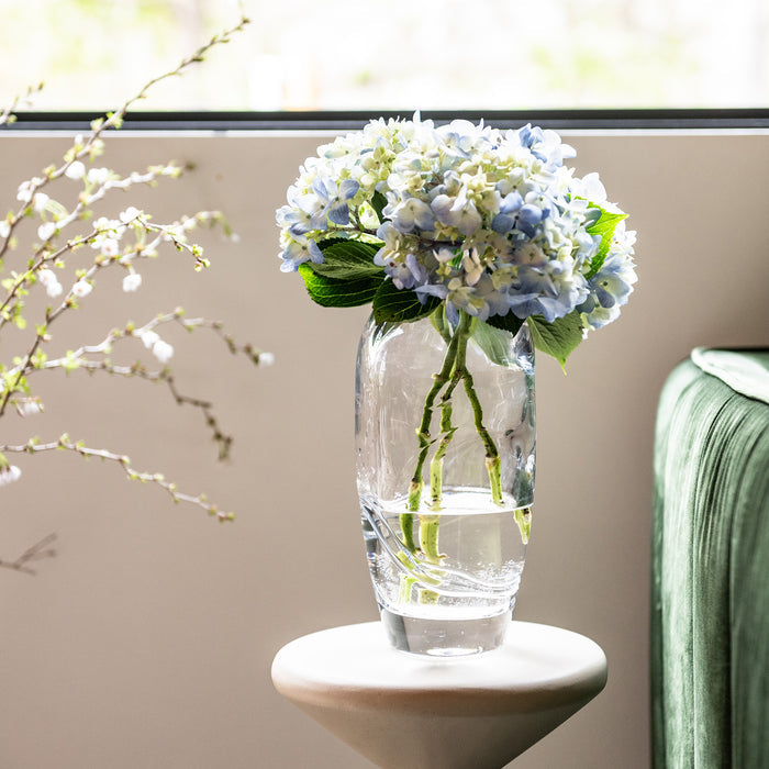 Clear glass vase with blue and white hydrangea flowers and green stems in water.
