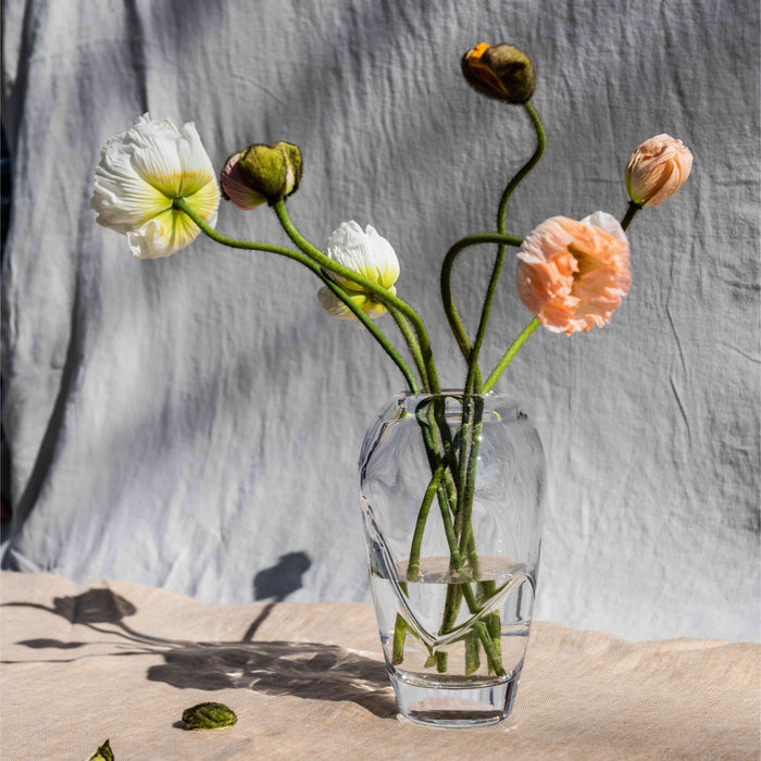 Clear glass vase with white and peach flowers and green stems arranged in water against a gray linen backdrop.