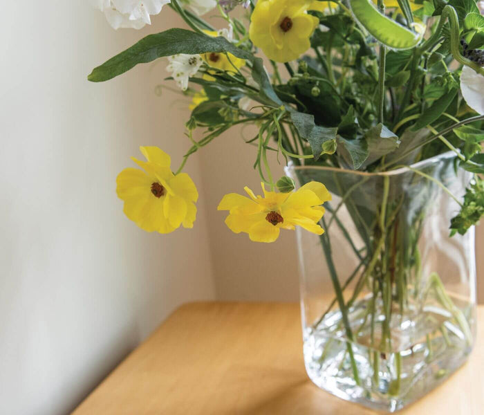 Clear glass vase filled with yellow and white flowers and green stems on a wooden surface.