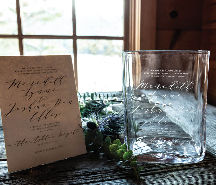 Clear glass rectangular vase with etched script text displayed next to wedding invitation and greenery.