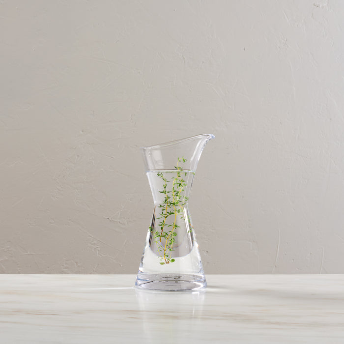 Clear glass vase with hourglass shape and flared rim, filled with water and small green plant sprigs.