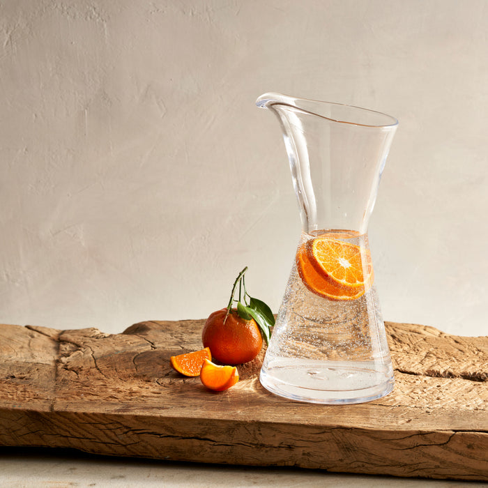 Clear glass pitcher with tapered shape and handle, filled with water and orange slice, positioned on weathered wood with fres