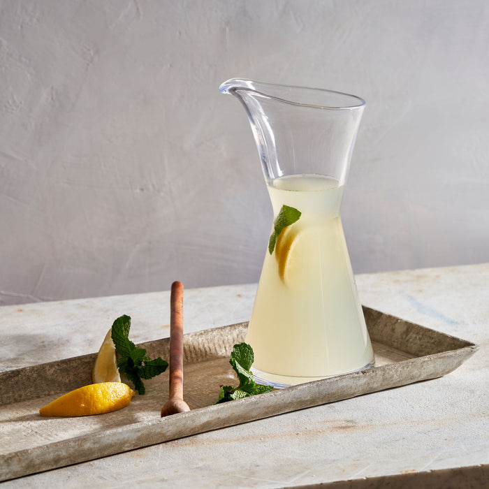 Clear glass pitcher filled with pale yellow liquid, lemon slice, and mint sprig inside, displayed on concrete tray with lemon