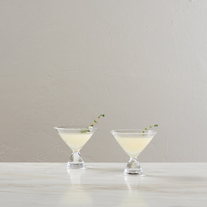 Two clear stemless martini glasses filled with pale cocktail, each garnished with herbs.