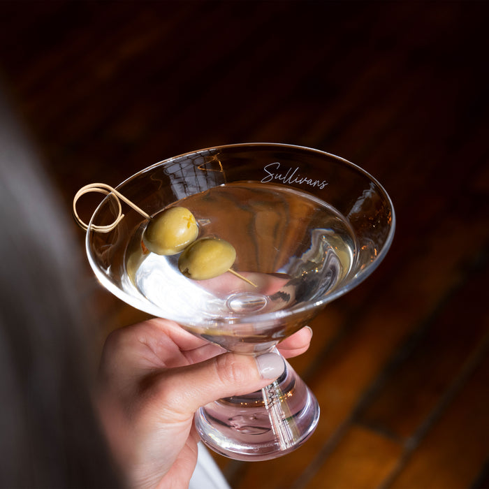 Hand holding a martini glass filled with clear liquid and two green olives on a pick, with "Sullivans" engraved on the rim.