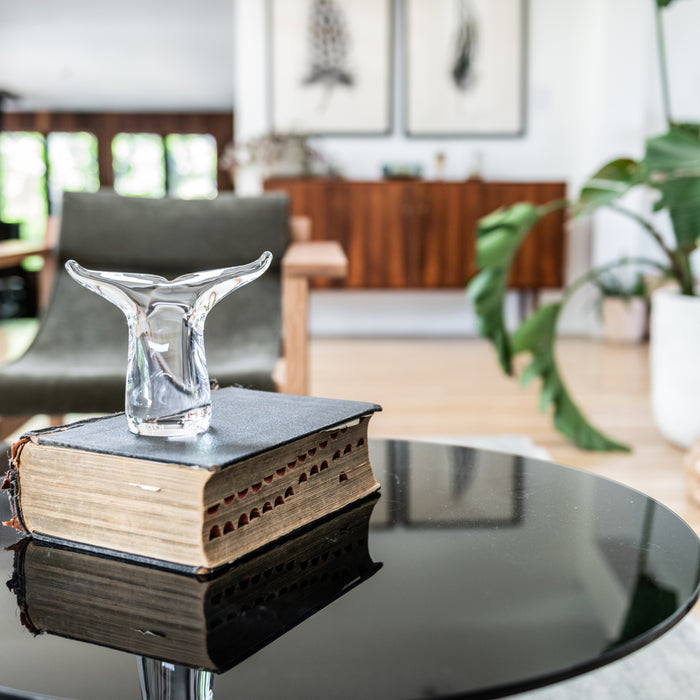 Clear glass whale tail figurine displayed on stacked books on a round black table in a modern living room.