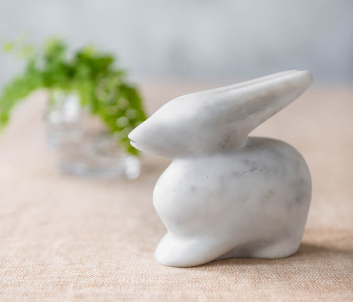 White marble rabbit figurine with pointed ears, displayed on beige surface with gift box in background.