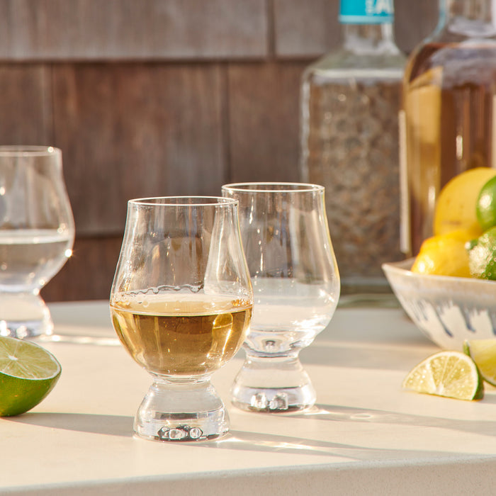 Two clear tasting glasses, one filled with amber liquid, displayed on a countertop with whiskey bottles and citrus fruits in
