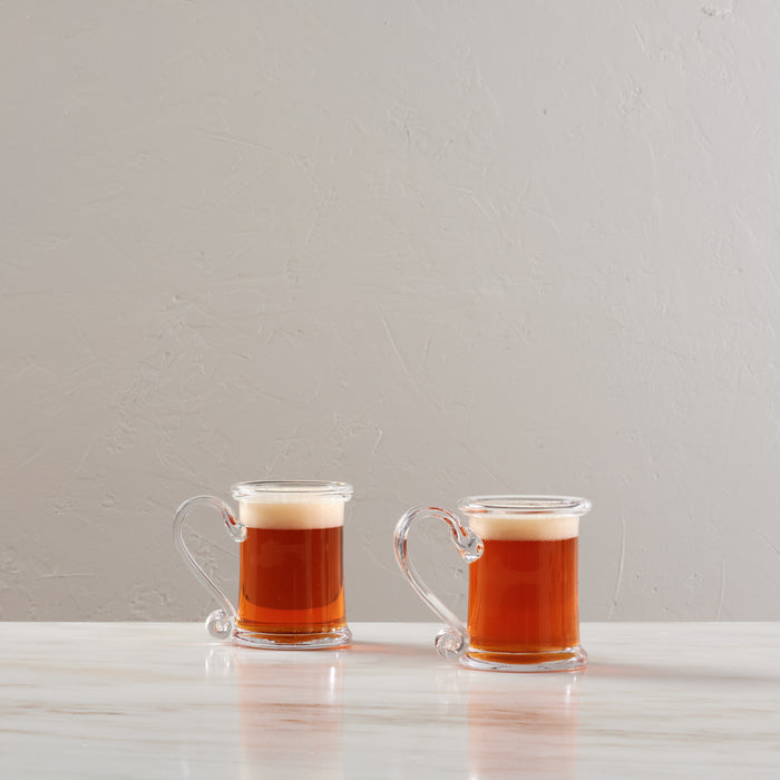 Two clear glass tankards with handles filled with amber liquid, shown against a white textured background.