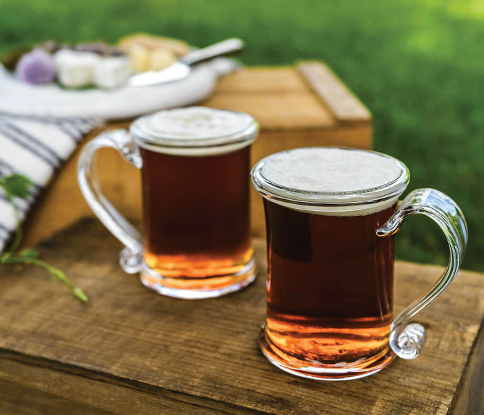 Two glass tankards with metal lids filled with dark beverage, displayed on a wooden board outdoors.