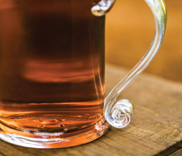 Clear glass tankard with handle filled with amber liquid, featuring decorative thumb rest detail.