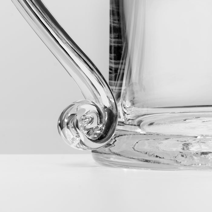 Close-up of a clear glass showing a curved glass handle with a decorative curled base and a thick textured glass body on a light gray background.