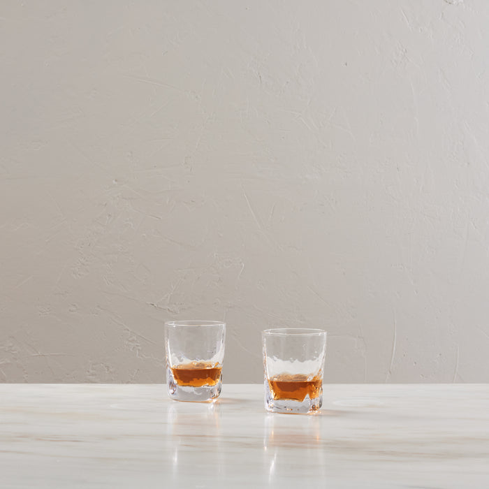 Two glasses filled with bourbon whiskey on a white surface against a textured beige wall.