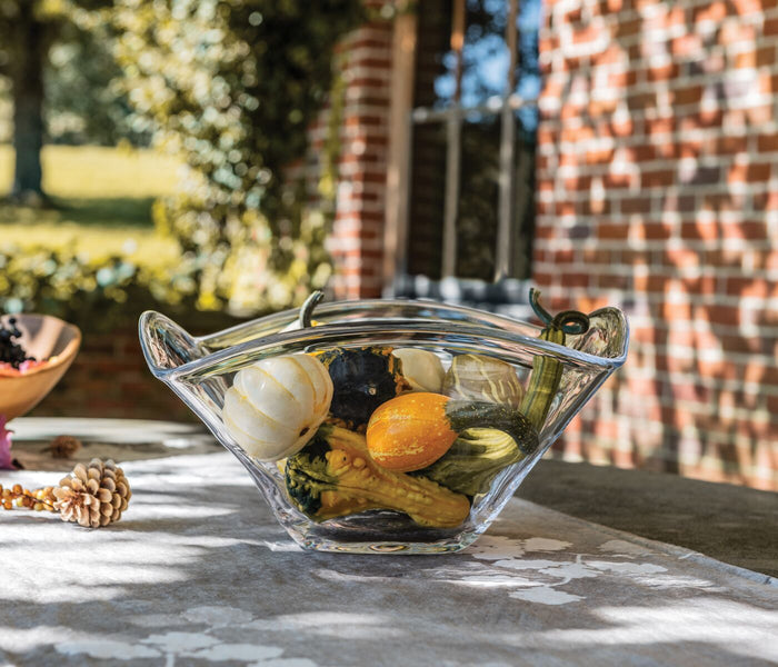 Clear glass bowl with wavy rim edges containing decorative gourds and vegetables, displayed outdoors on stone surface.