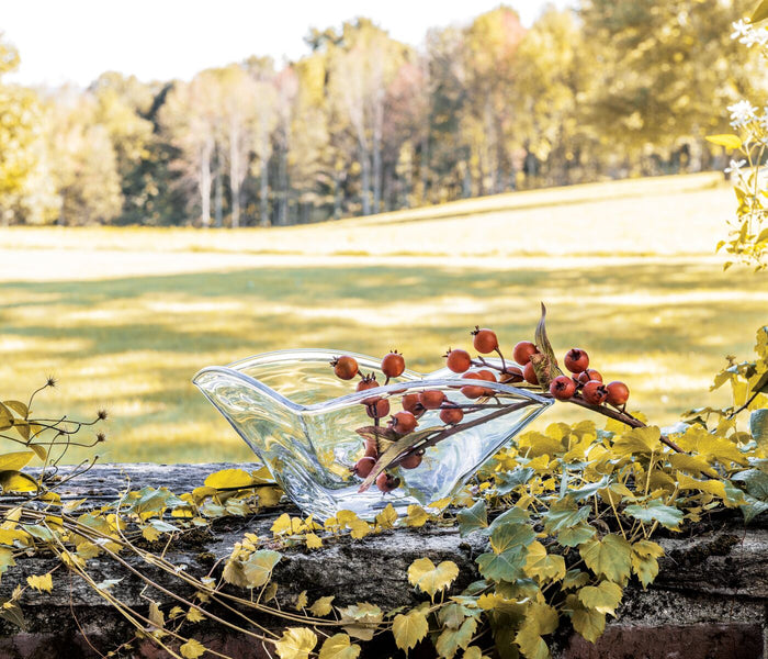 Clear glass bowl with curved rim containing a branch of red berries, displayed outdoors among autumn foliage.
