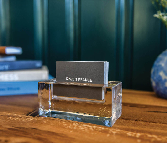 Clear glass cardholder with gray card slot displaying "SIMON PEARCE" branding on wooden desk.