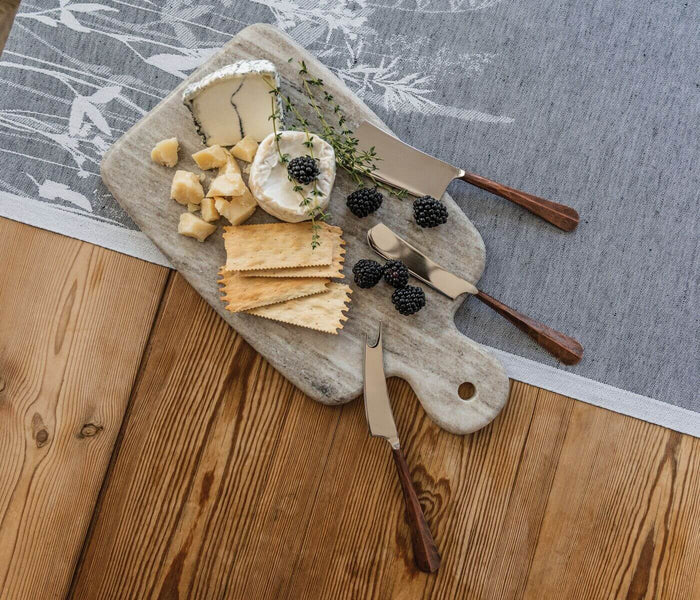 Three copper-handled cheese knives with wooden handles arranged on a gray marble board with assorted cheeses and berries.