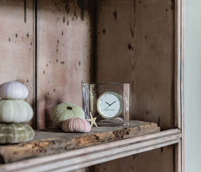 Square glass clock with white face and black hands displayed on wooden shelf with decorative sea urchins and driftwood.