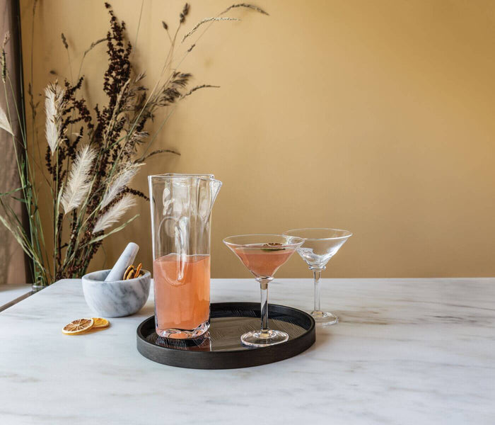 Clear glass carafe filled with coral-colored liquid, displayed on a black tray with two cocktail glasses.