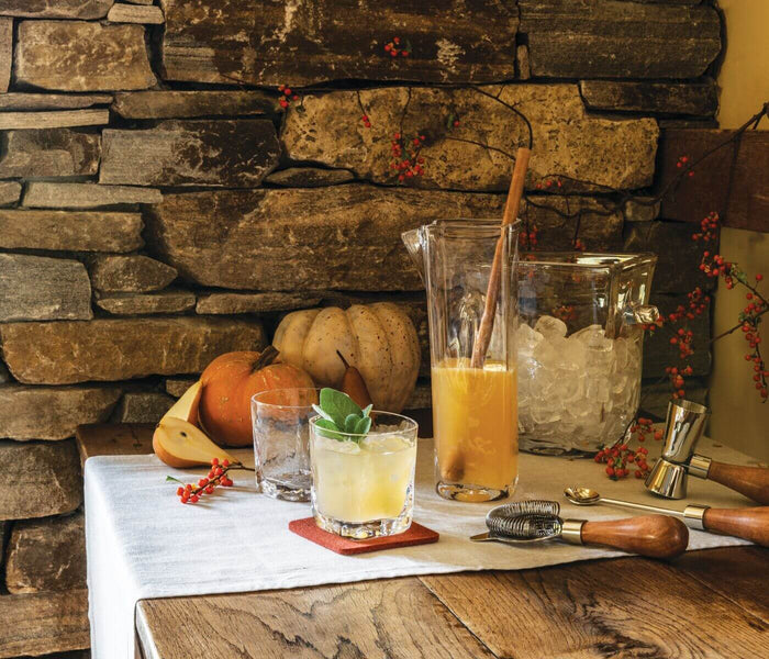 Clear glass carafe with wooden handle filled with yellow cocktail, displayed with mixed drinks and bar tools on white surface