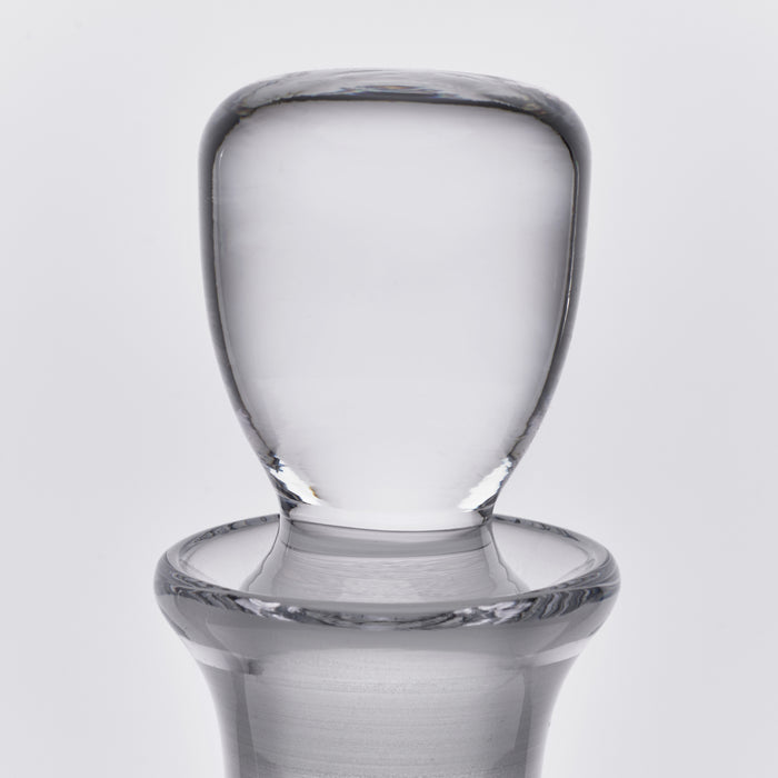 Clear glass decanter with rounded shoulders, cylindrical neck, and wide flat stopper on neutral background.