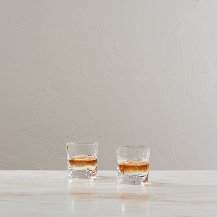 Two clear double old-fashioned glasses filled with amber liquid and ice cubes on white surface.
