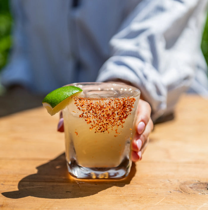 Clear glass tumbler filled with pale yellow cocktail, red spice rim, and lime wedge garnish, held by hand outdoors.