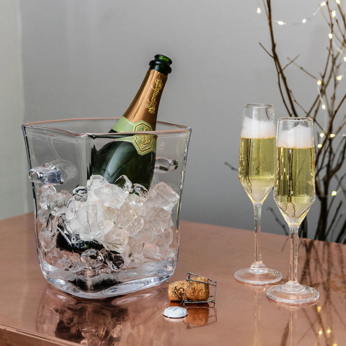 Clear glass ice bucket holding a champagne bottle surrounded by ice, with two filled champagne flutes beside it.
