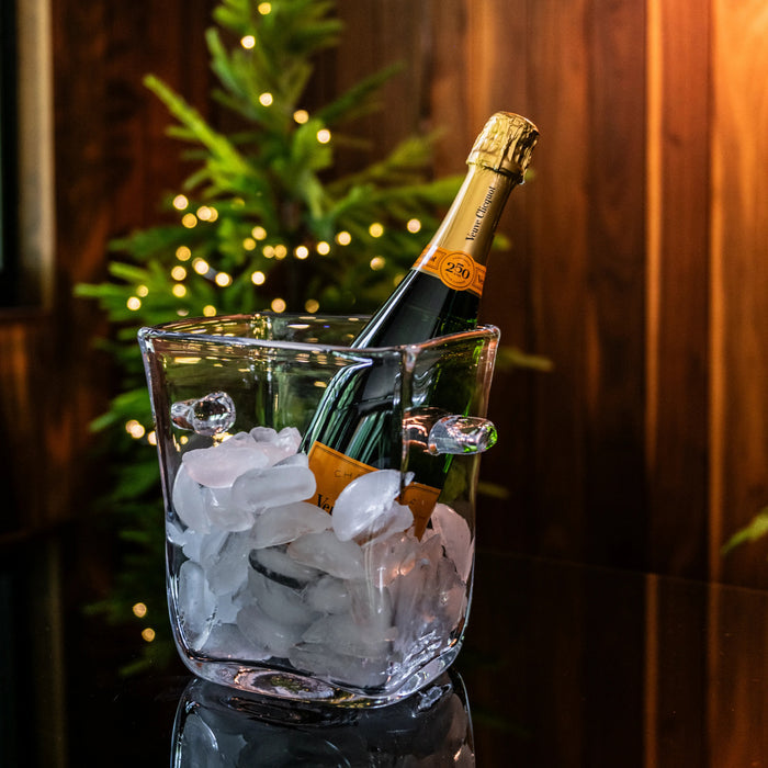 Champagne bottle in clear glass ice bucket filled with ice cubes, set against evergreen branches with warm lighting.