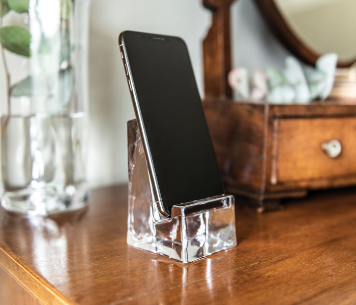 Clear glass phone holder with angled slot holding a smartphone upright on wooden surface.