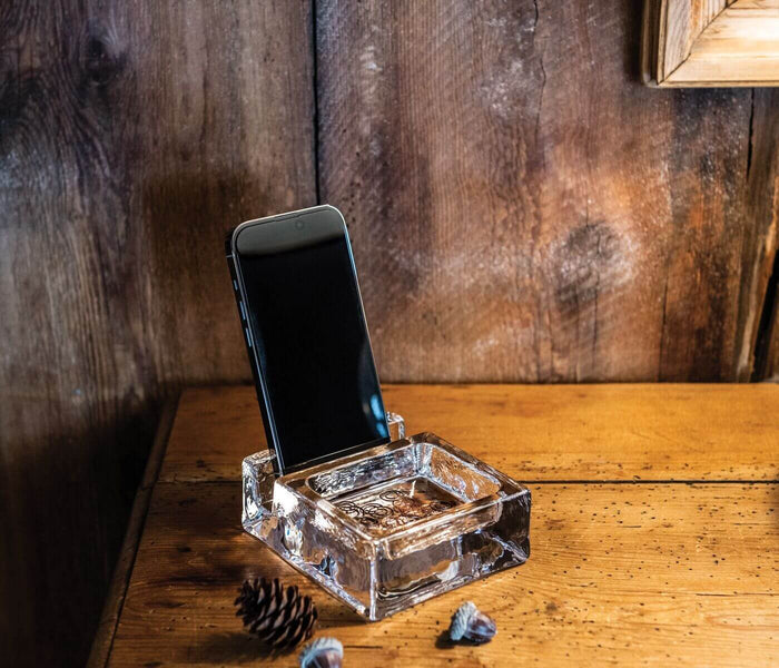 Clear glass phone stand holding a smartphone upright on a wooden surface with rustic wood paneling background.