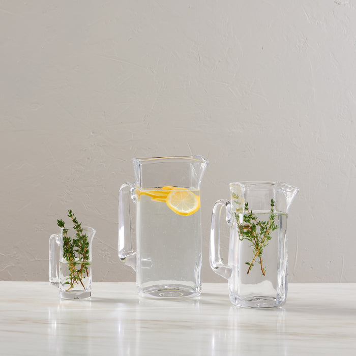 Clear glass pitcher with handle, containing water and lemon slice, displayed with two smaller matching glasses holding sprigs