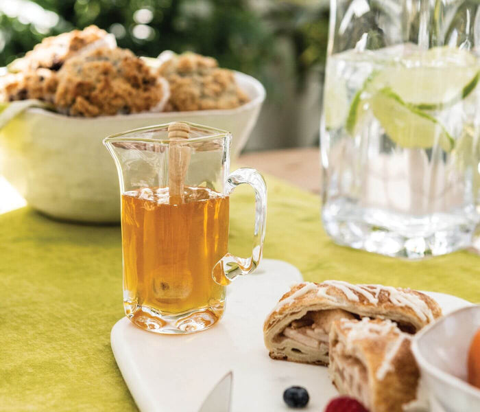 Clear glass pitcher filled with amber liquid and ice, with handle, surrounded by beverages and food on outdoor table.