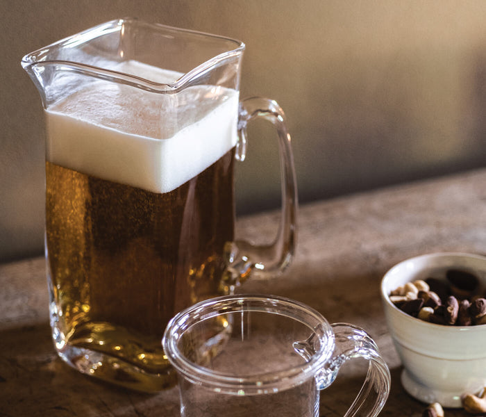 Clear glass pitcher filled with dark liquid and white foam, with matching cup and coffee beans nearby.